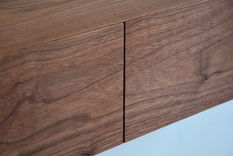 May include: A close-up view of two rectangular wooden drawer fronts featuring a rich, dark brown grain pattern. The panels are positioned side-by-side with a thin, vertical gap separating them, showcasing the natural texture and horizontal wood grain.