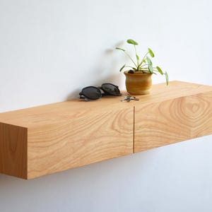 Op de afbeelding: Een lichtgekleurd houten zwevend rek aan een witte muur. Het heeft een kleine plant in een pot, een zonnebril en sleutels. De houtnerf is zichtbaar. Het rek is rechthoekig en ongeveer 120 cm lang.