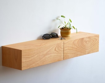 Puede incluir: Estantería flotante de madera clara montada en una pared blanca. Contiene una pequeña planta en maceta, gafas de sol y llaves. La veta de la madera es visible. La estantería es rectangular, de unos 120 cm de largo.