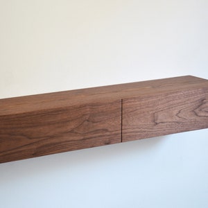May include: A rectangular floating wall shelf made of dark brown wood with a visible natural grain pattern. The shelf features two side-by-side drawer fronts that create a seamless, horizontal design mounted directly against a plain white wall.