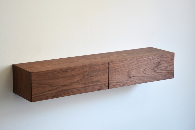 May include: A rectangular floating wall shelf made of dark brown wood with a visible natural grain pattern. The shelf features two side-by-side drawer fronts that create a seamless, horizontal design mounted directly against a plain white wall.