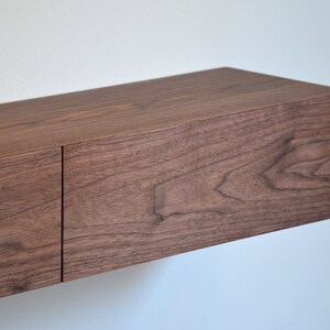 May include: A rectangular floating shelf made of dark brown wood with a visible natural grain pattern. The shelf features a smooth, flat top surface and a front panel divided into two sections by a vertical seam, creating a clean, linear appearance.