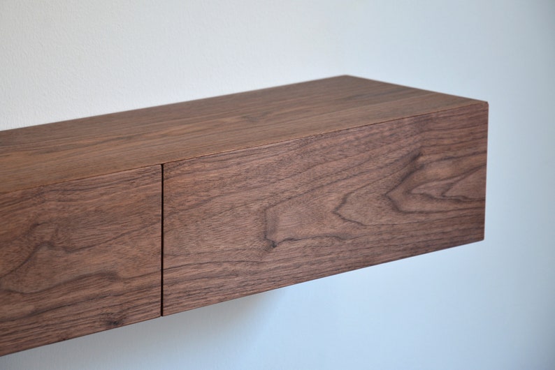 May include: A rectangular floating shelf made of dark brown wood with a visible natural grain pattern. The shelf features a smooth, flat top surface and a front panel divided into two sections by a vertical seam, creating a clean, linear appearance.