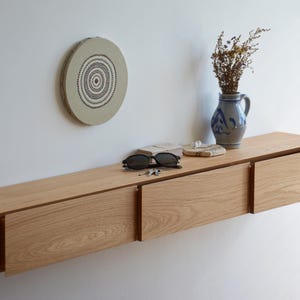 Puede incluir: Estante flotante de roble claro con tres cajones, montado en una pared blanca. El estante contiene un jarrón de cerámica azul y blanco con flores secas, gafas de sol y una decoración de pared redonda.