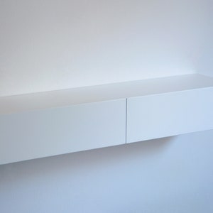 May include: A white floating shelf with two drawers. The shelf is mounted on a white wall.