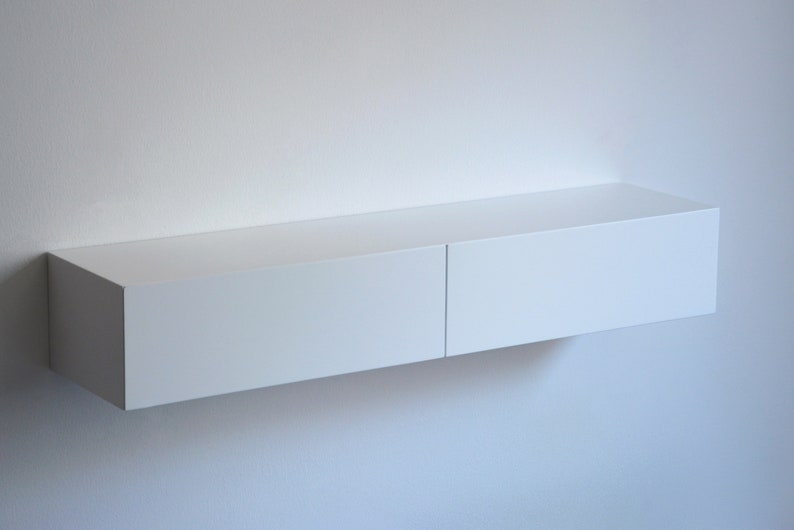 May include: A white floating shelf with two drawers. The shelf is mounted on a white wall.