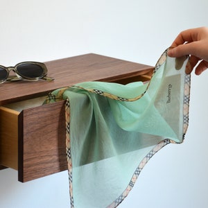 May include: A light mint green handkerchief with a beige, black, and red plaid border and the text Burberry printed on the fabric. A pair of dark-rimmed sunglasses rests on a wooden drawer unit next to the folded fabric accessory.
