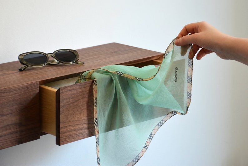 May include: A light mint green handkerchief with a beige, black, and red plaid border and the text Burberry printed on the fabric. A pair of dark-rimmed sunglasses rests on a wooden drawer unit next to the folded fabric accessory.