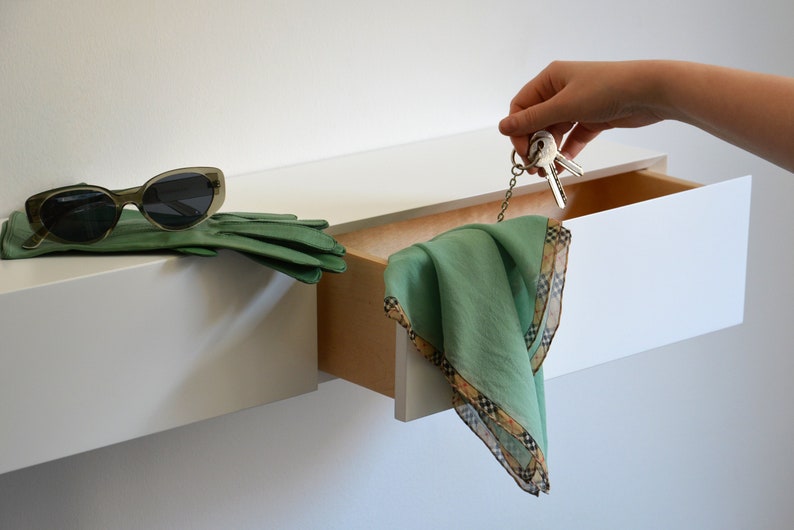 May include: A white floating shelf with a drawer that is partially open. A green scarf hangs out of the drawer, and a set of keys is attached to the scarf. A pair of sunglasses and green leather gloves are on the shelf.