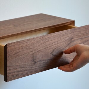 May include: A wall-mounted floating drawer unit crafted from dark brown walnut wood with a visible natural grain pattern. The rectangular drawer is partially extended, revealing a lighter-toned interior wood construction and a smooth front panel.