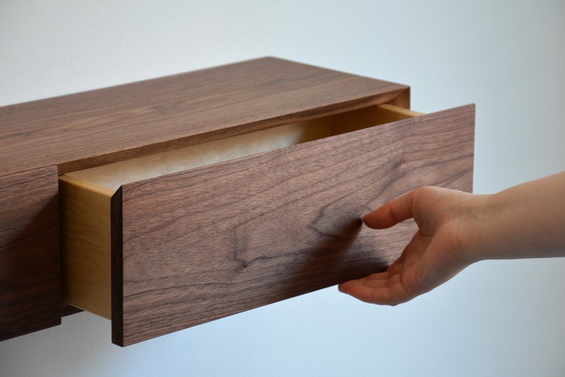 May include: A wall-mounted floating drawer unit crafted from dark brown walnut wood with a visible natural grain pattern. The rectangular drawer is partially extended, revealing a lighter-toned interior wood construction and a smooth front panel.