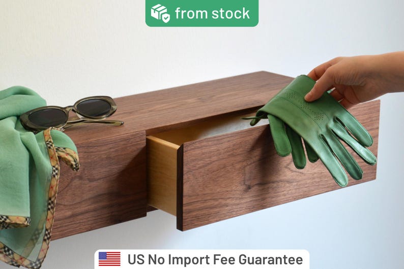 May include: A floating wall-mounted wooden drawer unit with a dark brown wood grain finish. A pair of green leather gloves, a light green scarf, and dark-rimmed sunglasses are placed on the surface of the drawer unit.
