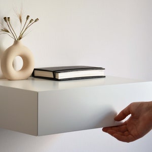 May include: A white floating shelf with a drawer that is being opened by a hand. A black notebook and a white ceramic vase with dried flowers are on the shelf.