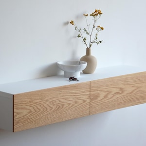 Op de afbeelding: Een zwevend houten dressoir met een wit blad en twee lades. Een kleine beige vaas met gele bloemen, een witte kom en sleutels staan op het dressoir. Het dressoir is aan een witte muur bevestigd.