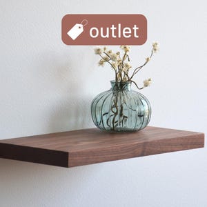 May include: A dark brown floating shelf against a white wall. A clear, ribbed glass vase with dried flowers sits on the shelf. A brown tag with the word "outlet" is in the top center.