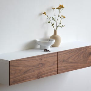 Op de afbeelding: Een modern zwevend rek met een wit bovenblad en een walnoothouten voorkant. Een kleine beige vaas met gele bloemen, een witte kom en sleutels staan op het rek. Het rek is aan een witte muur bevestigd.