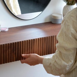 May include: A wooden vanity with a fluted drawer and a round mirror above. The vanity is mounted to a white wall. A person is opening the drawer.