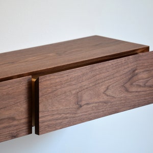May include: A wall-mounted floating shelf featuring two drawers constructed from dark brown walnut wood with a visible natural grain pattern. The drawers are positioned side-by-side with a horizontal orientation and a smooth, flat front surface.