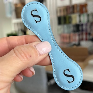 May include: A light blue, elongated, oval-shaped magnetic clip with rounded ends. The item features white stitched edges and a black letter S printed on each end. The surface has a smooth texture, and the overall design is symmetrical and flat.