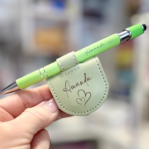 May include: A light green pen with a matching holder. The pen has a silver tip and accents, and the holder is a light green leather with the name "Amanda" and two hearts. The pen also has the name "Amanda" and a heart.