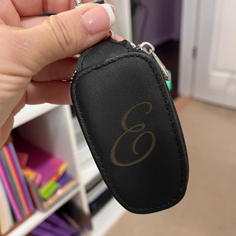 Puede incluir: Llavero de cuero negro con una letra "E" dorada en relieve en la parte delantera.