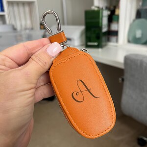 Puede incluir: Llavero de cuero naranja con un gancho plateado y una cremallera plateada. El llavero tiene la letra "A" grabada en la parte delantera.