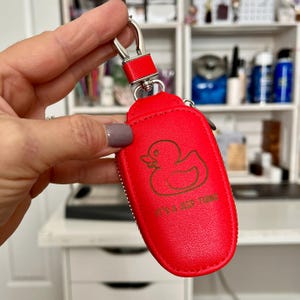 Puede incluir: Un llavero de cuero rojo brillante con un anillo de metal plateado y un dise&ntilde;o de pato de goma. El llavero tiene el texto "It's a Keep Thing" impreso en &eacute;l.