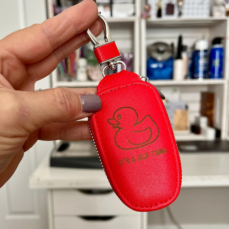 Puede incluir: Un llavero de cuero rojo brillante con un anillo de metal plateado y un dise&ntilde;o de pato de goma. El llavero tiene el texto "It's a Keep Thing" impreso en &eacute;l.