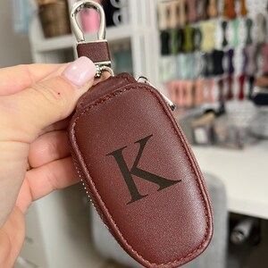 Puede incluir: Un llavero de cuero marr&oacute;n con un lazo plateado y una cremallera. El llavero tiene la letra "K" grabada en la parte delantera.