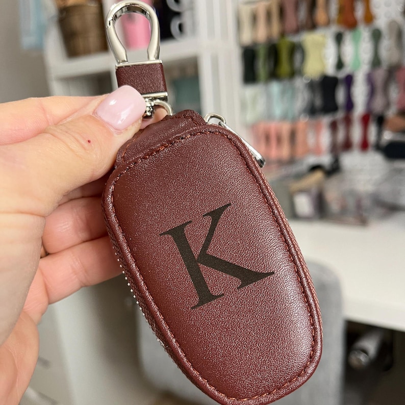 Puede incluir: Un llavero de cuero marr&oacute;n con un lazo plateado y una cremallera. El llavero tiene la letra "K" grabada en la parte delantera.