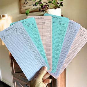 May include: A hand holds a fan of five pastel-colored reading log cards. Each card has a header that reads "Reading Log" and includes sections for book title, date, and rating. The cards are in shades of blue, pink, and green.