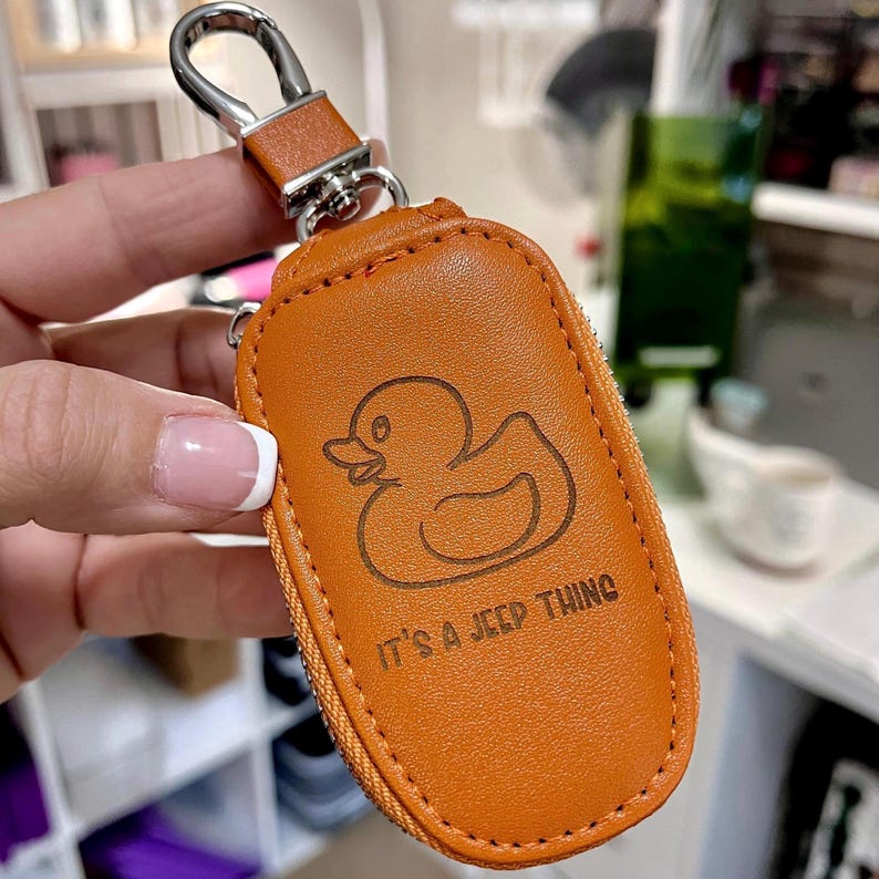 Puede incluir: Un llavero de cuero marr&oacute;n con un anillo de metal y un dise&ntilde;o de pato de goma. El llavero tiene el texto "It's a Jeep thing" grabado.