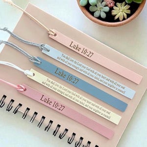 May include: Four leather bookmarks in pastel colors: pink, blue, and cream. Each bookmark has the text "Luke 10:27" and a Bible verse. The bookmarks are attached to a spiral notebook with a beige cord.