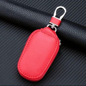 Puede incluir: Porta llaves de cuero rojo con un gancho de metal plateado. El porta llaves tiene una cremallera.