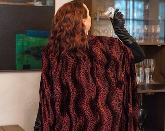 Könnte beinhalten: Eine Frau trägt einen roten und schwarzen Leopardenfellmantel mit langen schwarzen Handschuhen. Sie steht vor einem Schrank aus Holz mit Glastüren und hält ein Glas in ihrer rechten Hand.