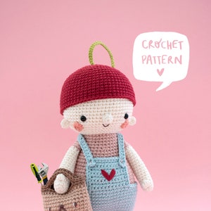 Puede incluir: Muñeca de ganchillo hecha a mano con un peto azul, una gorra roja y botas marrones. La muñeca sostiene una pequeña bolsa marrón con herramientas. El texto "CROCHET PATTERN" está en un bocadillo. El fondo es rosa.