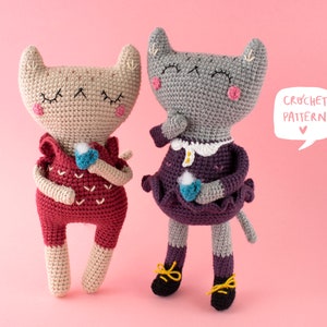 May include: Two crocheted cat dolls, one tan and one gray, stand side-by-side against a pink backdrop. The tan cat wears a red outfit and holds a blue object. The gray cat wears a purple dress and black shoes. A speech bubble says "CROCHET PATTERN".