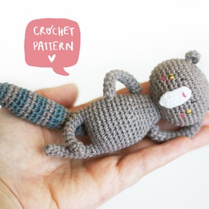 Puede incluir: Un juguete de ardilla gris tejido a ganchillo con una cola a rayas azules y grises, tumbado boca arriba. La ardilla tiene un hocico blanco, detalles rosas y ojos amarillos. La imagen incluye una burbuja de diálogo rosa con las palabras "CROCHET PATTERN".