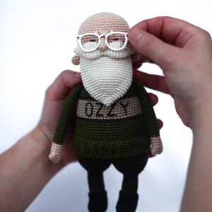May include: A handmade crochet doll of an elderly man with a white beard and glasses. The doll wears a green sweater with the name "OZZY" on it. The doll's skin is tan, and it has black pants and shoes. The doll is held by two hands.