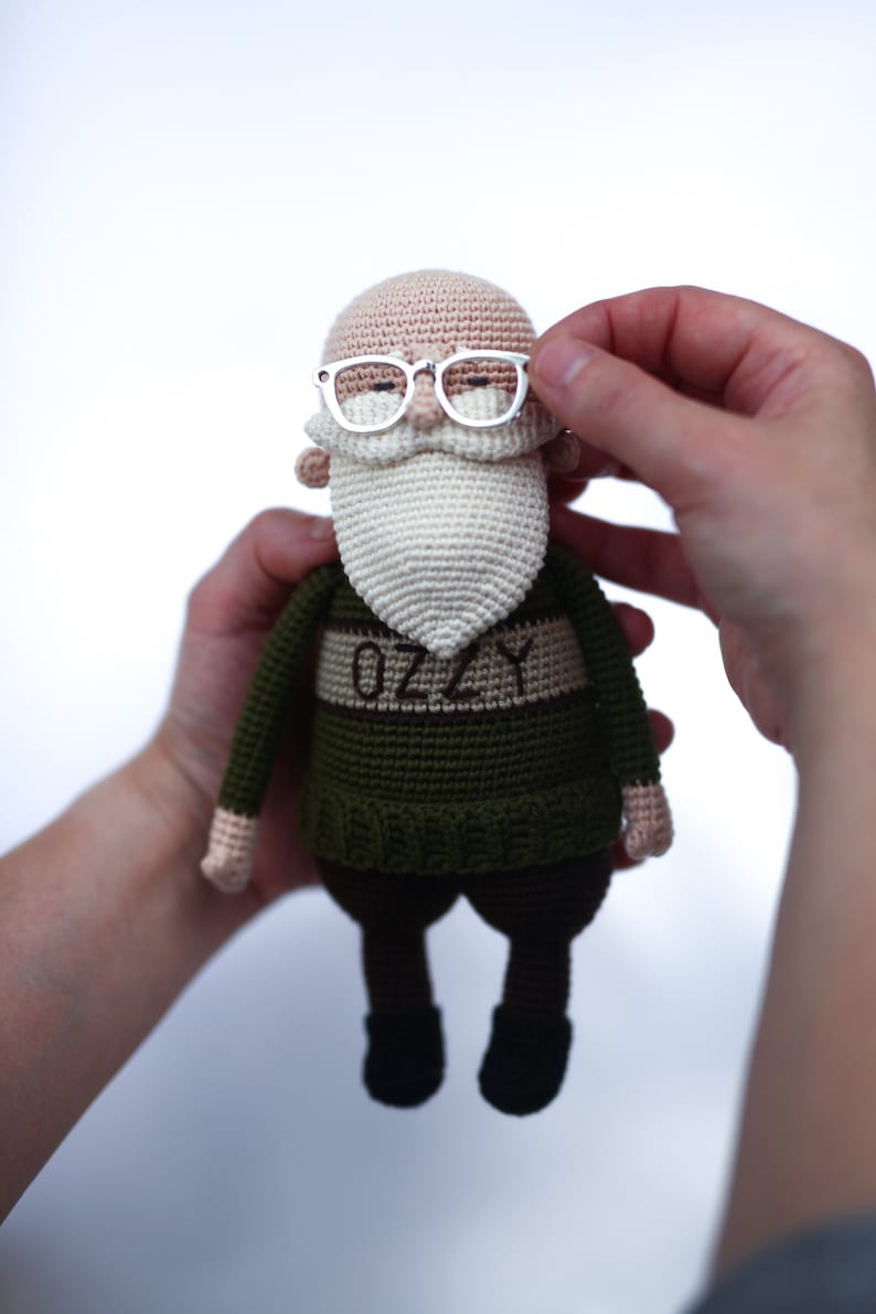 May include: A handmade crochet doll of an elderly man with a white beard and glasses. The doll wears a green sweater with the name "OZZY" on it. The doll's skin is tan, and it has black pants and shoes. The doll is held by two hands.
