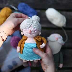 May include: Crocheted doll depicting an elderly woman with glasses, a bun, and a mustard-colored jacket over a blue dress. A second doll, a crocheted man with a beard, is also visible. Yarn in various colors is in the background.