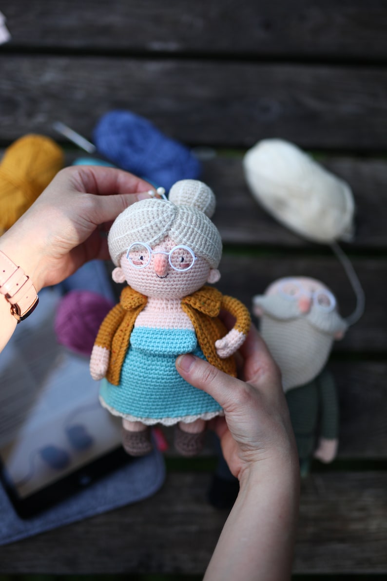 May include: Crocheted doll depicting an elderly woman with glasses, a bun, and a mustard-colored jacket over a blue dress. A second doll, a crocheted man with a beard, is also visible. Yarn in various colors is in the background.