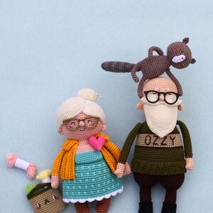 May include: Crocheted dolls of an elderly couple holding hands, with a squirrel on the man's head. The woman wears glasses, a yellow jacket, and a heart. The man has a white beard and glasses. A small basket and yarn are nearby.