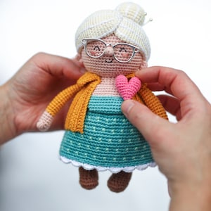May include: A handmade crochet doll depicting a smiling elderly woman with glasses, a yellow cardigan, and a teal dress. The doll has a white bun and is holding a pink heart. The doll is approximately 6 inches tall.