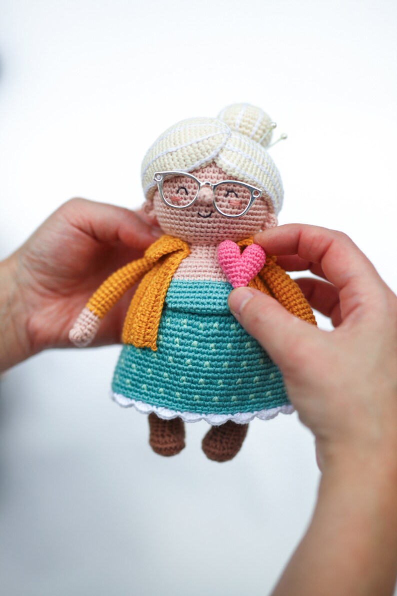 May include: A handmade crochet doll depicting a smiling elderly woman with glasses, a yellow cardigan, and a teal dress. The doll has a white bun and is holding a pink heart. The doll is approximately 6 inches tall.