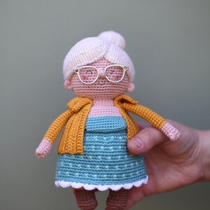 May include: A handmade crochet doll depicting an elderly woman. The doll wears a blue dress with white polka dots, a yellow cardigan, and a cream-colored bun. It has gold-colored glasses and brown shoes. The doll is held by a hand.