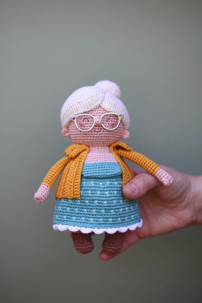 May include: A handmade crochet doll depicting an elderly woman. The doll wears a blue dress with white polka dots, a yellow cardigan, and a cream-colored bun. It has gold-colored glasses and brown shoes. The doll is held by a hand.