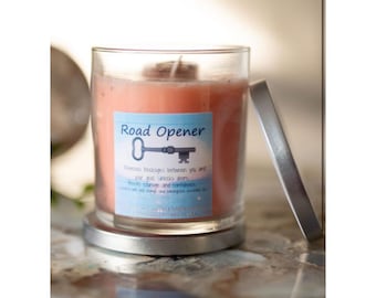 May include: A glass candle jar with a silver lid. The candle is a peach color and has a label that reads "Road Opener" with a key graphic and the text "Removes blockage between you and your goals. Unlocks doors.  Develops courage and confidence.  With citrus and mahogany essential oils."
