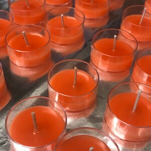 May include: A collection of orange-colored tea light candles in clear plastic holders. Each candle has a central wick. The candles are arranged on a reflective silver surface, creating a visually appealing display. The candles are a warm, inviting color.