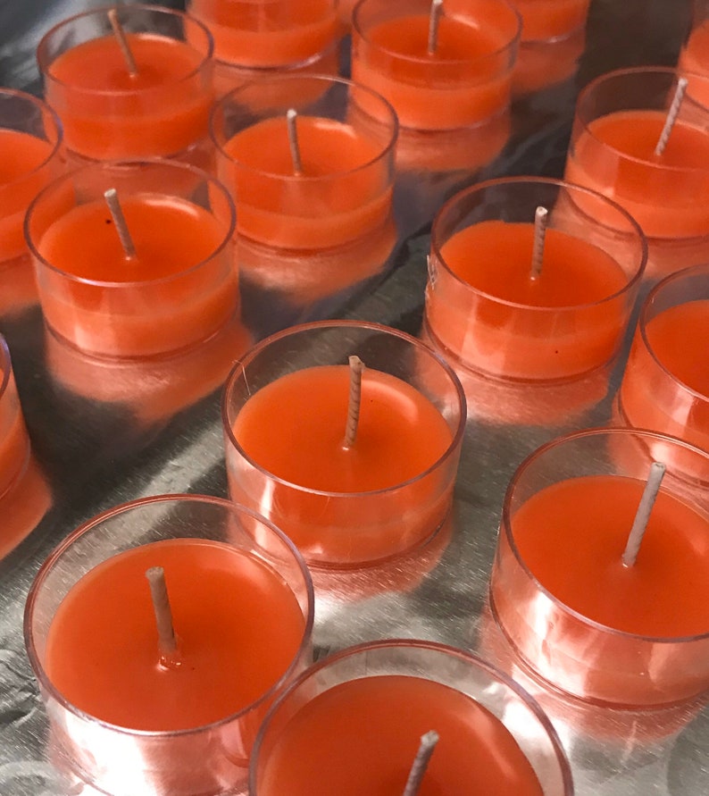 May include: A collection of orange-colored tea light candles in clear plastic holders. Each candle has a central wick. The candles are arranged on a reflective silver surface, creating a visually appealing display. The candles are a warm, inviting color.